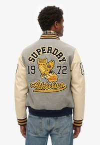 Grijze wollen varsityjack met crèmekleurige mouwen, voorzien van geborduurde gele graphics, waaronder "SUPERDRY" en "Athletics", gestreepte geribde manchetten.