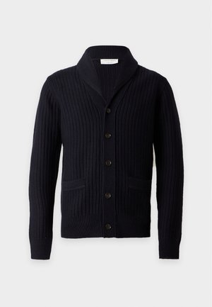 Homme portant un cardigan noir boutonné, un t-shirt blanc, un pantalon cargo gris et des chaussures noires, debout devant un fond blanc uni.