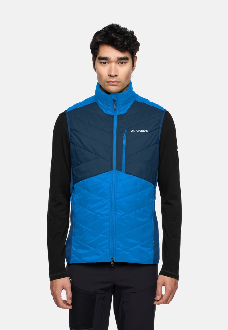 Blauwe geïsoleerde vest met een hoge kraag, diagonaal gestikt patroon, borstzak en ritssluitingen, gedragen over een zwart longsleeve shirt.