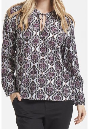 Frau trägt eine langärmelige Bluse mit lila-schwarzem Blumenmuster und Ausschnitt mit Schlitz, kombiniert mit schwarzer Hose.