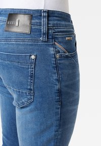 Närbild av blå denimjeans på en mannekäng, med bakfickor, bältesöglor, metallnitar och en svart varumärkesetikett med texten "MAVI ULTRAMOVE".