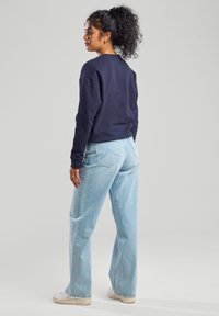 Marineblauwe sweatshirt met ronde halslijn, gecombineerd met lichtblauwe wijde jeans. Model staat met de rug naar de camera, waardoor de relaxte pasvorm en casual stijl zichtbaar zijn.