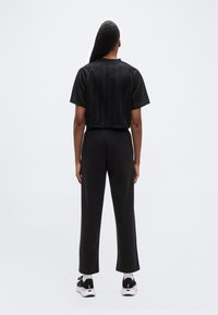 Άτομο που στέκεται με την πλάτη, φοράει κοντομάνικη μαύρη κορυφή, μαύρο παντελόνι, λευκές κάλτσες και μαύρα-λευκά αθλητικά παπούτσια, απλό φόντο.