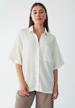 Camicia bianca, con texture, a maniche corte e colletto, dotata di taschino sul petto, abbinata a pantaloni beige.