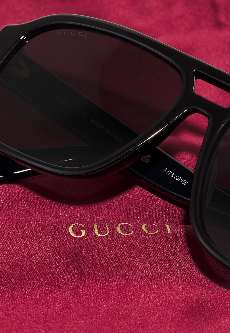 Lunettes de soleil noires Gucci avec des verres sombres, présentant une forme rectangulaire audacieuse. Exposées sur un tissu rouge avec des détails de logo doré.