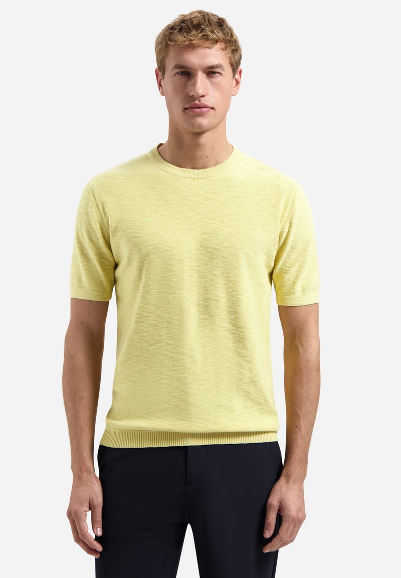 Man draagt een lichtgeel, gebreid, kort mouwtje trui en donkere broek, staande tegen een effen witte achtergrond.