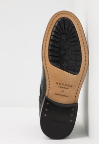 Černá kožená podrážka s vroubkovanou gumou pro lepší trakci, s hnědým okrajem a reliéfním logem. Obsahuje malé kovové kloboučky blízko paty.