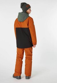 Orangene und schwarze wasserdichte Jacke mit einer grünen Kapuze, kombiniert mit orangen Schneehosen und einer grauen Mütze. Robustes Material und lässiger Schnitt.