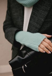 Gants sans doigts en tricot vert menthe avec une texture côtelée portés par une main reposant sur un sac noir élégant avec un détail de boucle argentée.