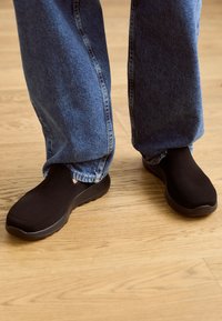 Czarne buty wsuwane z teksturowaną siateczką i wyściełaną podeszwą, w połączeniu z luźnymi niebieskimi dżinsami na drewnianej podłodze.