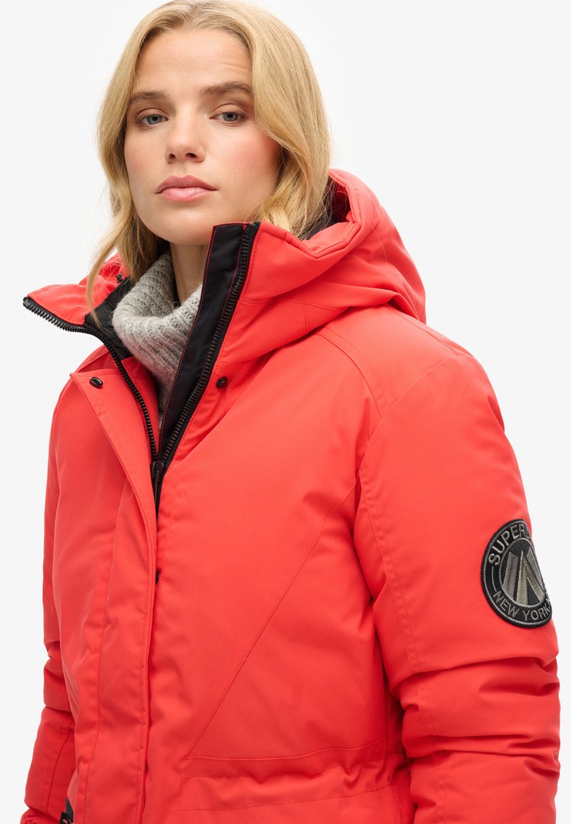 Rote isolierte Jacke mit Kapuze, schwarzem Reißverschluss und einem strukturierten grauen Schal. Verfügt über ein rundes Abzeichen am linken Ärmel.