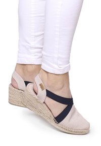 Sandal wedge yang menampilkan bagian atas kanvas beige dengan tali silang navy, midsole jute bergaya espadrille, menampilkan finish yang bercorak.