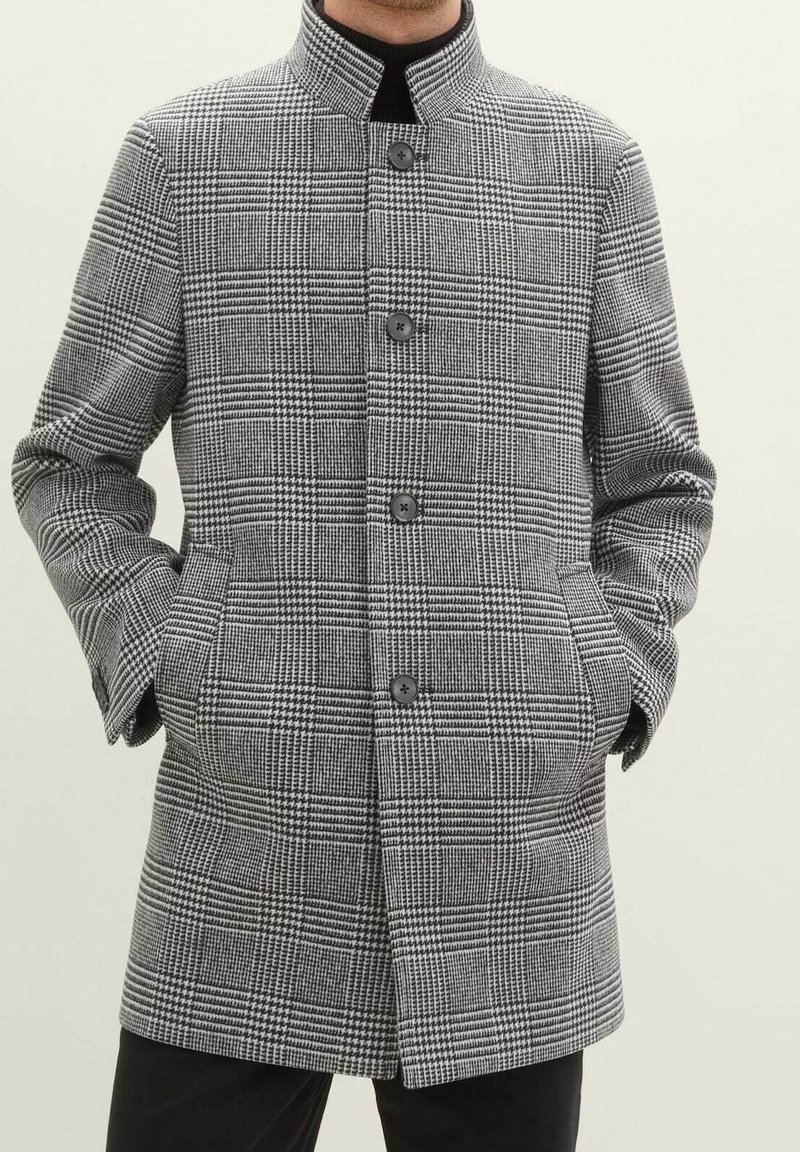 Płaszcz w czarno-białą kratę z wysokim kołnierzem, zapinany na guziki z przodu, z bocznymi kieszeniami. Wykonany z teksturowanej tkaniny.