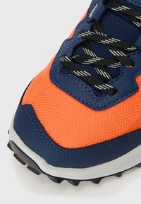 Pomarańczowe i granatowe buty sportowe z teksturowaną siateczką, czarno-białymi sznurówkami w paski oraz wytrzymałą podeszwą z bieżnikiem.