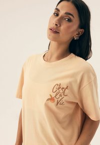 Różowa bawełniana koszulka z graficznym nadrukiem z napisem "C'est la Vie" oraz pomarańczowym akcentem w lewej części klatki piersiowej.