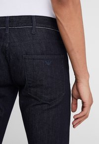 Jean en denim bleu foncé avec une poche arrière ornée d'un logo discret et des surpiqûres contrastantes. La coupe est slim avec un design de jambe fuselée.