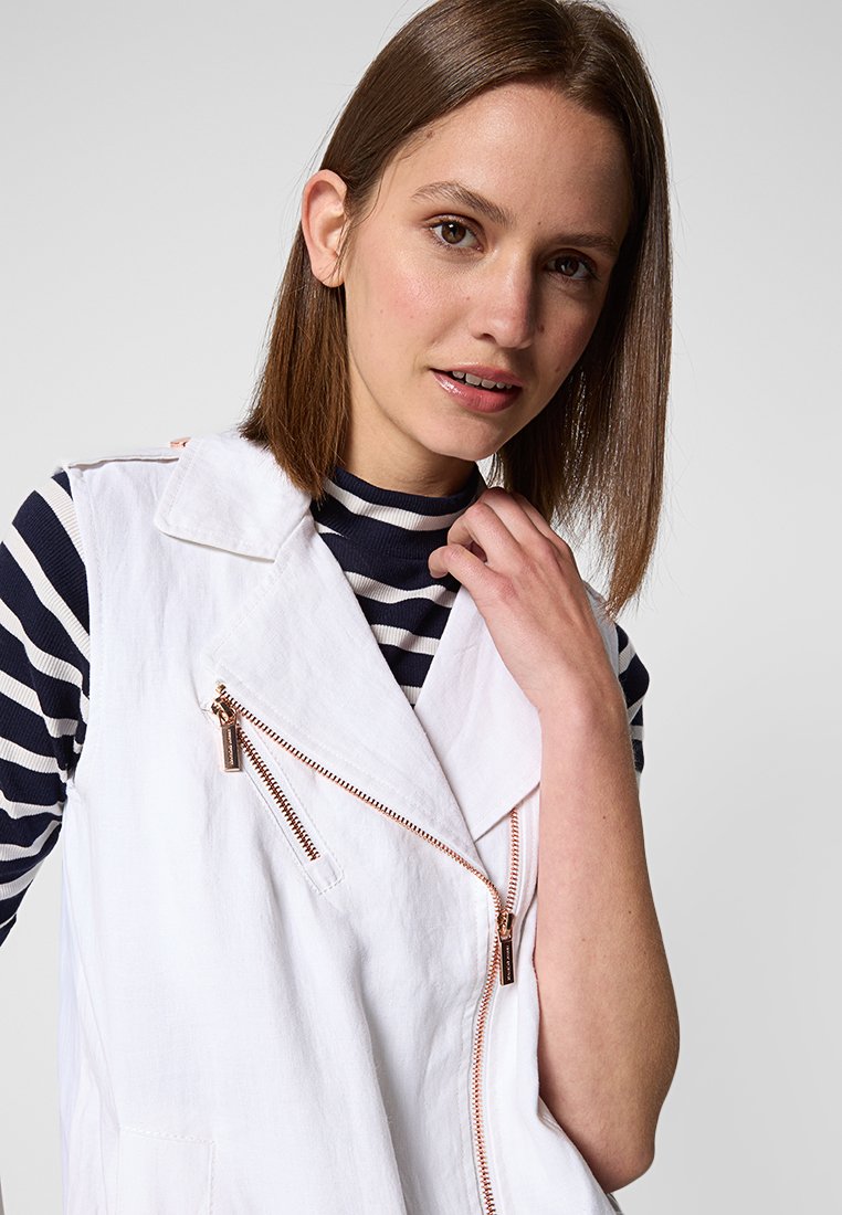 Gilet bianco senza maniche con una zip diagonale e accessori in oro rosa. Sotto, un dolcevita a righe blu navy e bianche. Texture del tessuto liscia.