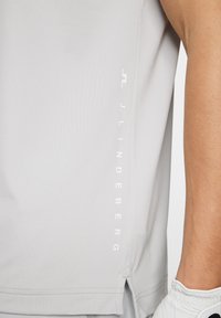 Light gray athletic tank top made from smooth fabric, featuring a side slit and "J.LINDEBERG" logo in white along the edge.