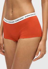 Γυναίκα με πορτοκαλί εσώρουχο τύπου boyshort με λευκή ζώνη μέσης με σήμανση "BJÖRN BORG" στέκεται μπροστά σε απλό φόντο.