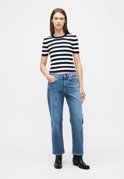 Γυναίκα με κοντομάνικη ριγέ μαύρο-άσπρη μπλούζα, μπλε τζιν παντελόνι με ευθεία γραμμή και μαύρα μποτάκια αστραγάλου, στέκεται μπροστά σε απλό φόντο.