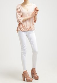 Vaaleanpunainen V-neulostyylinen paita, jossa on 3/4-hihat ja solmupisto vyötäröllä, yhdistettynä valkoisiin tiukkoihin farkkuihin ja nudenvärisiin korkokenkiin.