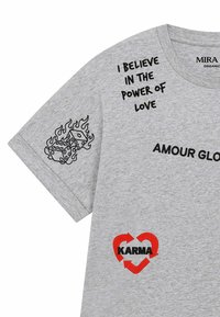 T-shirt gris avec texte noir « I believe in the power of love », « AMOUR GLO », motif de dés en flammes sur la manche, et cœur rouge avec flèches et « KARMA ».