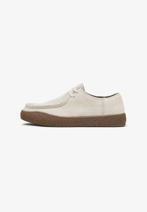 Zapato de ante beige con punta redondeada, dos ojales para cordones y suela de goma marrón texturizada. Diseño sencillo, perfil plano.