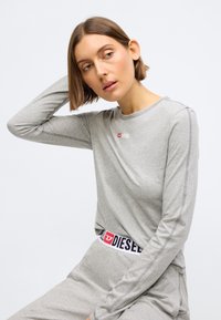 Svijetlosivi dugi rukav majica sa zaobljenim izrezom, s malim crvenim logotipom na prsima i pojasom u crnoj i crvenoj boji sa brandom. Mekana tekstura.