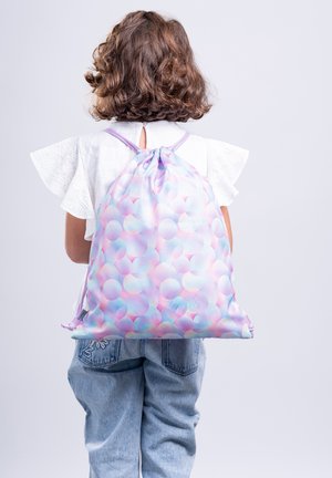 Bambino con capelli castani ricci, indossa una camicetta bianca e jeans, porta uno zaino con coulisse color pastello con motivo circolare sulla schiena.