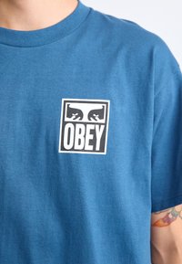 Blå bomull t-skjorte med en hvit grafikk som har teksten "OBEY" og stiliserte ansikter innrammet i en firkant på venstre brystområde.