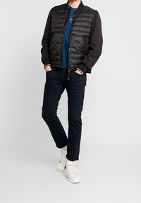Zwarte pufferjack met een doorgestikt ontwerp, blauwe shirt eronder, donkere jeans en witte sneakers. Het jack heeft ribgebreide manchetten en een kraag.