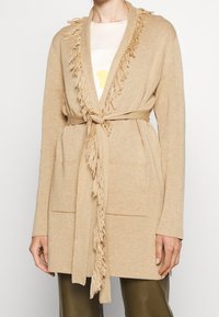 Cardigan beige en tissu doux, avec un col à franges et des poches avant. Comprend une ceinture assortie pour la fermeture.