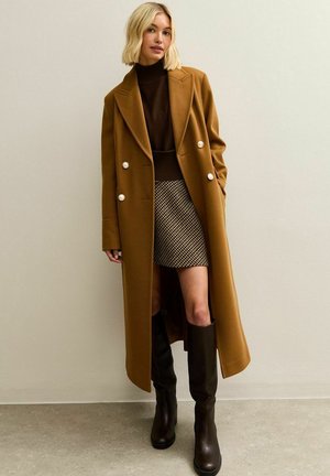 Mujer con un abrigo largo color camello con botones plateados, suéter de cuello alto marrón, falda de cuadros y botas negras hasta la rodilla, de pie contra una pared beige.