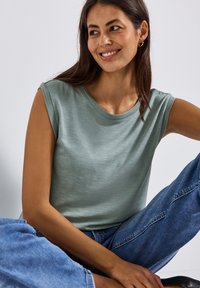 Jasnozielony, krótki top z okrągłym dekoltem i teksturowanym materiałem. Noszony z niebieskimi dżinsami, prezentując luźny krój i swobodny styl.