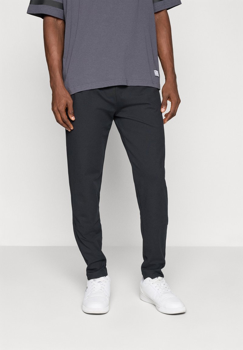 Dunkelgraue Herrenhose mit schlanker Passform, aus leichtem Stoff. Kombiniert mit einem dunkelgrauen Kurzarmhemd und weißen Sneakern.