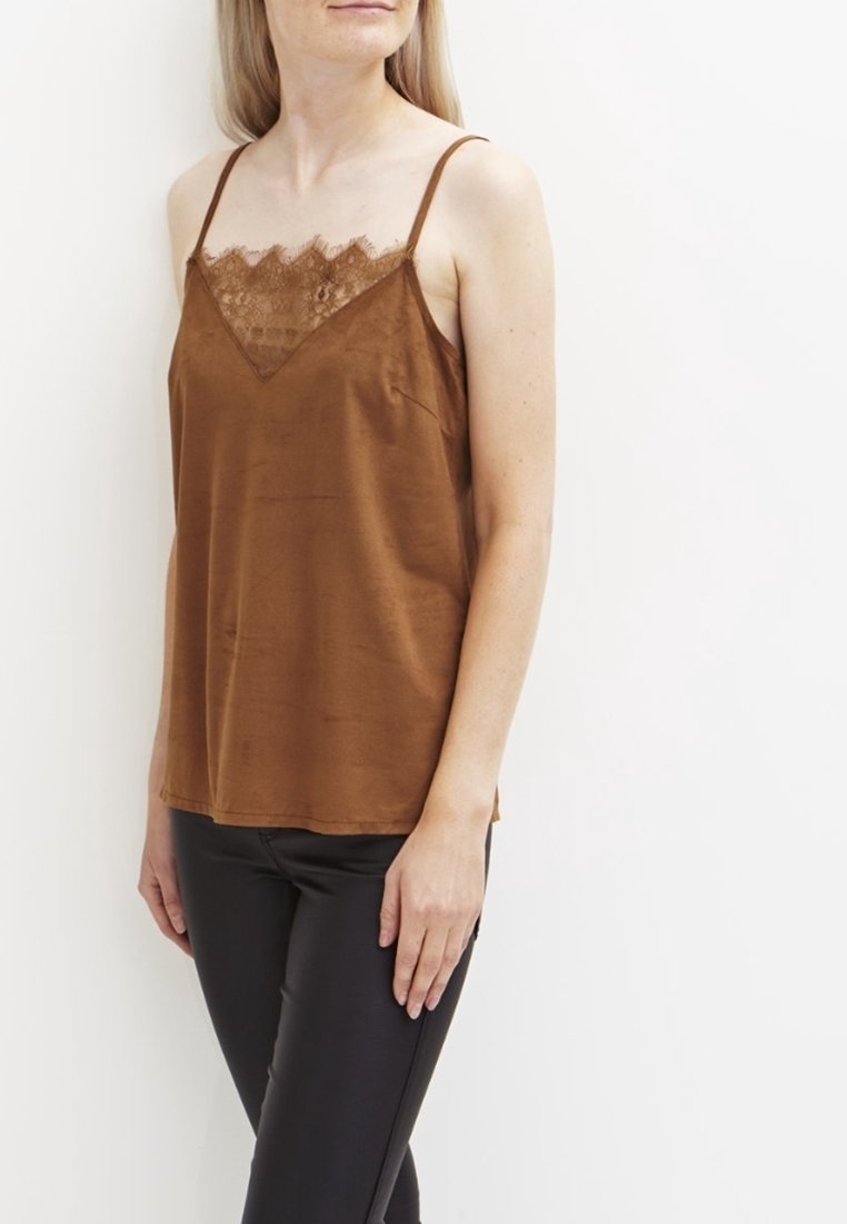 Femme portant un haut sans manches marron orné de dentelle et un pantalon noir, debout sur un fond blanc uni.