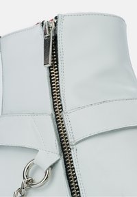 White leather boot featuring a prominent silver zipper, circular metal accent, and detailed stitching. Textured material and sleek design.