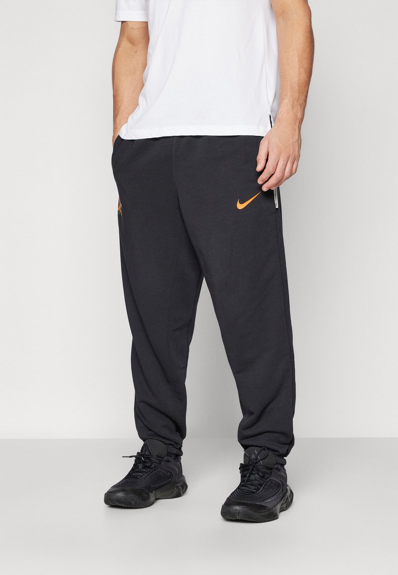Černé joggingové kalhoty s oranžovým logem Nike, vyrobené z měkkého materiálu, s elastickými manžetami a postranními kapsami, nošené s černými teniskami.