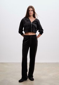 Kvinde står indendørs iført en sort lynlås crop hoodie og matchende sorte drawstring bukser med sorte sneakers.