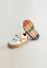 Witte canvas sneakers met oranje en blauwe accenten, dubbele klittenbandsluitingen en een geprofileerde rubberen zool met chevronstrepen in meerdere kleuren.