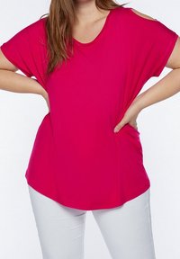 Woman wearing bright pink short-sleeve top with shoulder cutouts and white pants, hands resting on hips against plain background.