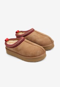 Brązowe zamszowe kapcie wsuwane z czerwonym haftowanym obramowaniem, miękką wyściółką i gumową podeszwą. Posiadają pętelkę ułatwiającą zakładanie.