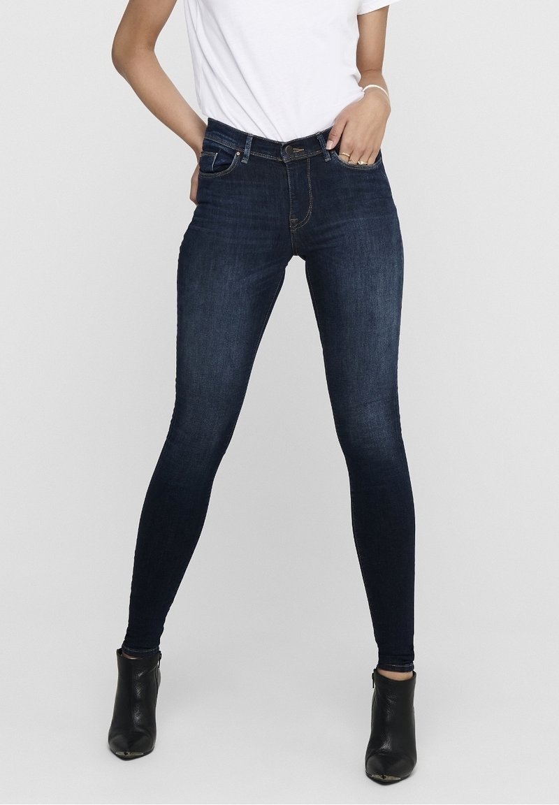 Skinny jeans in dark blue denim, featuring a classic five-pocket design and slight whiskering at the hips. Worn with black ankle boots.