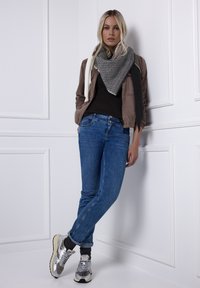 Bruine jas, zwart topje, blauwe spijkerbroek, grijze sjaal met structuur, zilveren sneakers met beige accenten. Model leunt tegen een witte paneelmuur.