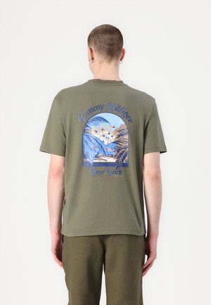 Olive zeleno bombažno majico s velikim modrim in kremnim grafičnim motivom s palmami ter napisom "Tommy Hilfiger New York". Kratki rokavi, okrogla ovratnika.