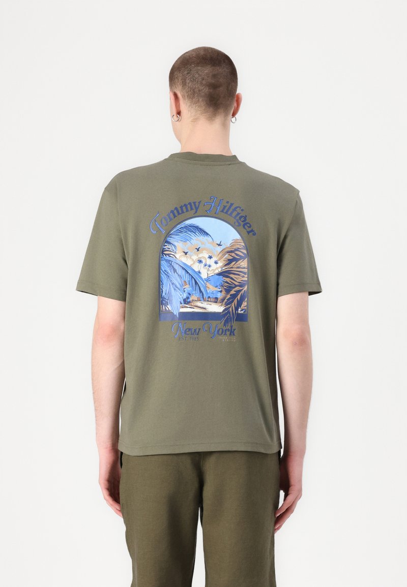 Camiseta de algodón en verde oliva con un gran gráfico azul y crema que presenta palmeras y el texto "Tommy Hilfiger New York". Manga corta, cuello redondo.