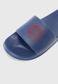 Blå sklidesandal laget av teksturert materiale med et preget rødt blomsterdesign på stroppen. Åpen tådesign med flate såler.