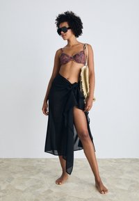 Dyreprint bikini topp, svart gjennomsiktig omslagskjørt med sidesløyfe og store solbriller. Teksturert overflate på steingulv.