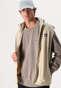 Beige fleece vest with zip closure, worn over a light brown sweatshirt. Grey cap. Both garments feature minimal branding and a relaxed fit.