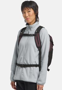 Une personne portant une veste en polaire gris clair zippée et un sac à dos violet et noir avec des sangles de poitrine et de taille attachées.